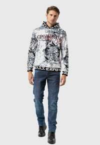 Sweat-shirt à capuche blanc avec des imprimés graphiques noirs et rouges, doté d'une capuche à cordon et d'un ourlet noir. Associe à un jean en denim bleu.