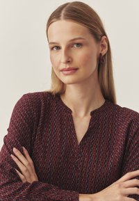 Burgundia pluus geomeetrilise mustriga punaste ja siniste linkidega, millel on lõikekaelus ja pikad varrukad, koos tumepunaste tilga kujuliste kõrvarõngastega.