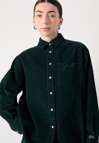 Camicia in corduroio verde scuro con colletto a camicia, taschino frontale e logo ricamato sulla manica. Presenta bottoni bianchi e una texture a coste.