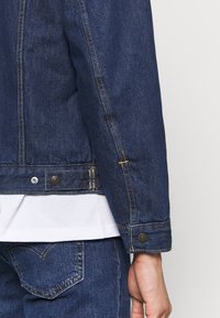 Dark blue denim jacket with contrasting stitching, snap buttons, and a structured design. Features a classic collar and straight hem.