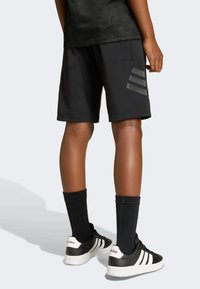 Crni atletski šorts s glatkom teksturom, s tri tamno sive pruge sa strane, uparen s crnim čarapama i crnim tenisicama.