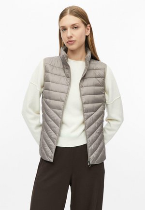 Junge Frau trägt eine hellgraue gesteppte Weste über einem weißen Pullover mit braunen Hosen und steht mit den Händen hinter dem Rücken.
