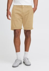 Shorts din bumbac bej cu manșete răsucite, având închidere cu nasture în față, loop-uri pentru curea și buzunare laterale, asortați cu adidași gri și șosete albe.