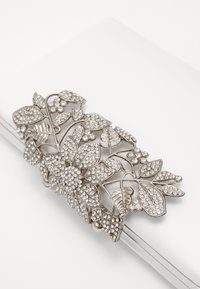 Stříbrná květinová spona do vlasů zdobená malými průhlednými kamínky spočívající na bílé zrcadlové ploše.