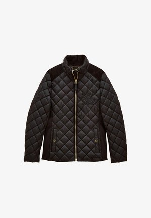 Frau mit lockigen Haaren, die eine schwarze gesteppte Jacke über einem Pullover mit Ski-Fuchs-Motiv trägt, kombiniert mit weißen Hosen vor neutralem Hintergrund.