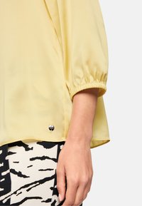 Yellow blouse with gathered sleeves, lightweight fabric, and a small black logo accent near the hem, paired with patterned pants.