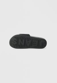 Black slide sandal sole facing up with textured tread and raised text "JEANS" on the bottom.