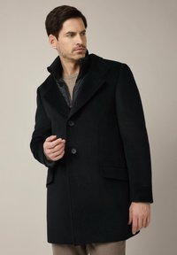 Czarny płaszcz z mieszanki wełny z klapami, zapinany na jeden guzik, z dwoma guzikami i bocznymi kieszeniami, nałożony na wewnętrzną kurtkę zapinaną na zamek.