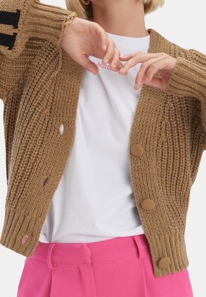 Personne portant un cardigan en grosse maille marron sur un t-shirt blanc et un pantalon rose vif, tenant les mains près de la poitrine.