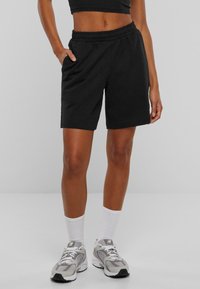 Femme portant un short à taille élastique noir, des chaussettes blanches montant jusqu'au mollet et des chaussures de sport grises, debout devant un fond uni gris clair.