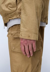 Ανδρικό χέρι ακουμπισμένο στην τσέπη παντελονιού cargo σε ανοιχτό καφέ χρώμα, φοράει αντίστοιχο ανοιχτό καφέ μπουφάν με ορατό πλεκτό γκρι μανσετέ από μέσα.