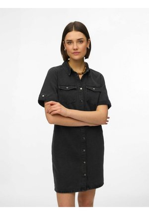 Jonge vrouw met kort bruin haar, gekleed in een zwarte spijkerjurk met knopen en een kralenketting, staande met gekruiste armen tegen een witte achtergrond.