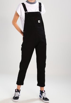 Sorte bomuldsoverall med justerbare skulderstropper, sidesknapper og en brystlomme, kombineret med en hvid t-shirt og blå høje sneakers.