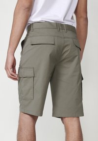 Uomo che indossa pantaloni cargo verde oliva e una maglietta bianca, visto da dietro, con tasche a patta sui lati e sul retro dei pantaloni.