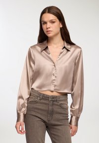 Camicia in satin corta di un color taupe chiaro con chiusura a bottoni, maniche lunghe e colletto classico, abbinata a jeans scuri a vita alta.
