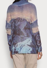 Personne portant un pull côtelé avec un motif de paysage montagneux bleu et violet, associé à un pantalon beige clair, vu de derrière.