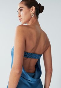 Satenasta modra obleka s strapless dizajnom, z nabrano hrbtom in nizkim izrezom, ki razkriva hrbet. Zlate uhane v obliki obroča dodajajo poudarke.