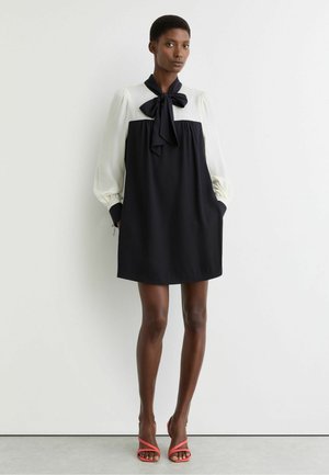 Žena stojí pri bielej stene, oblečená v čierno-bielych šatách s dlhým rukávom, s golierom s mašľou a koralovými sandálmi na podpätku.