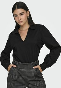 Blusa nera a maniche lunghe con scollatura a V e design con colletto, abbinata a pantaloni a quadri a vita alta con tasche frontali e dettagli con bottoni.