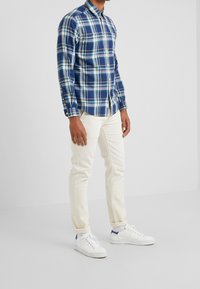 Chemise à carreaux bleue et verte avec boutons, manches longues, tissu en coton. Associée à un pantalon blanc ajusté et des baskets blanches avec des accents bleus.