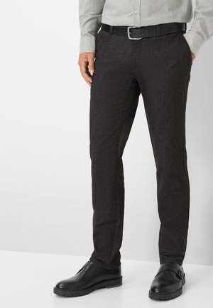 Mann trägt dunkelgraue, schmal geschnittene Hose, schwarzen Gürtel, schwarze Lederschuhe und ein hellgraues, langärmliges Hemd mit Knöpfen, steht mit den Händen in den Taschen.