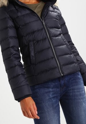 Marineblauer Pufferjacke mit einer Kunstfellkapuze, gestepptem Design, frontalem Reißverschluss und seitlichen Taschen. Über einem grauen Oberteil getragen und mit Jeans kombiniert.