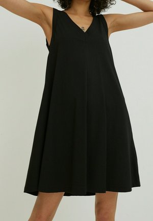 Kvinde iført en ærmeløs sort A-formet kjole, armene løftet, med synlig tatovering på øvre bryst mod en ensartet lys baggrund.