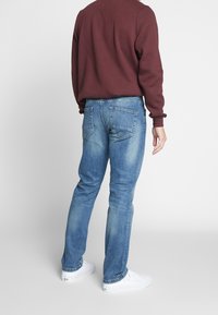 Donkerrode trui met structuur, blauwe denim jeans met een lichte wassing en twee achterzakken, gecombineerd met witte schoenen. Achteraanzicht van de outfit.