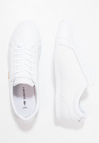 Bílé tenisky s hladkým syntetickým svrškem, zavazováním na tkaničky, strukturovanými bočními panely a podešví s logem, které mají minimalistický design.