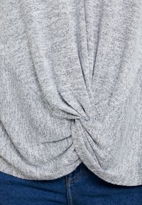 Haut en maille grise chinée avec un design torsadé à l'avant, une texture douce et une coupe décontractée, associé à un jean bleu.