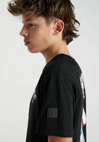 T-shirt noir en coton à manches courtes, présentant une impression graphique colorée au dos et un petit patch logo sur la manche.