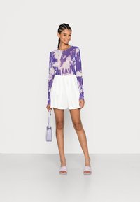 T-shirt de mangas longas em tie-dye roxo, saia plissada branca, bolsa lavanda e sandálias com o dedo do pé aberto, tudo numa base neutra.