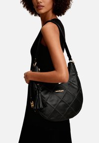 Donna con abito nero che porta una borsa a spalla in pelle nera trapuntata con nappina e dettaglio del logo dorato.