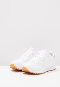 Vita sneakers med en slät läderyta, grå accenter, platta snören och en orange gummisula. Minimalistisk design med perforerade detaljer.