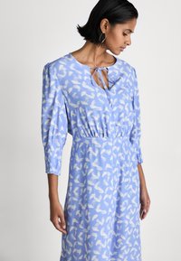 Rochie bleu deschis cu un model abstract alb, având un guler rotund, detaliu cu funda și mâneci trei sferturi cu manșete strânse.