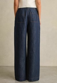 Spodnie dżinsowe o szerokich nogawkach w ciemnym indygo, z dwoma tylnymi kieszeniami i kontrastującym pomarańczowym przeszyciem, o gładkiej, teksturowanej powierzchni.