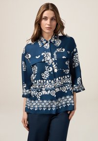 Blusa de tela suave con estampado floral azul, cuello tipo camisa, dos bolsillos frontales y mangas de 3/4.