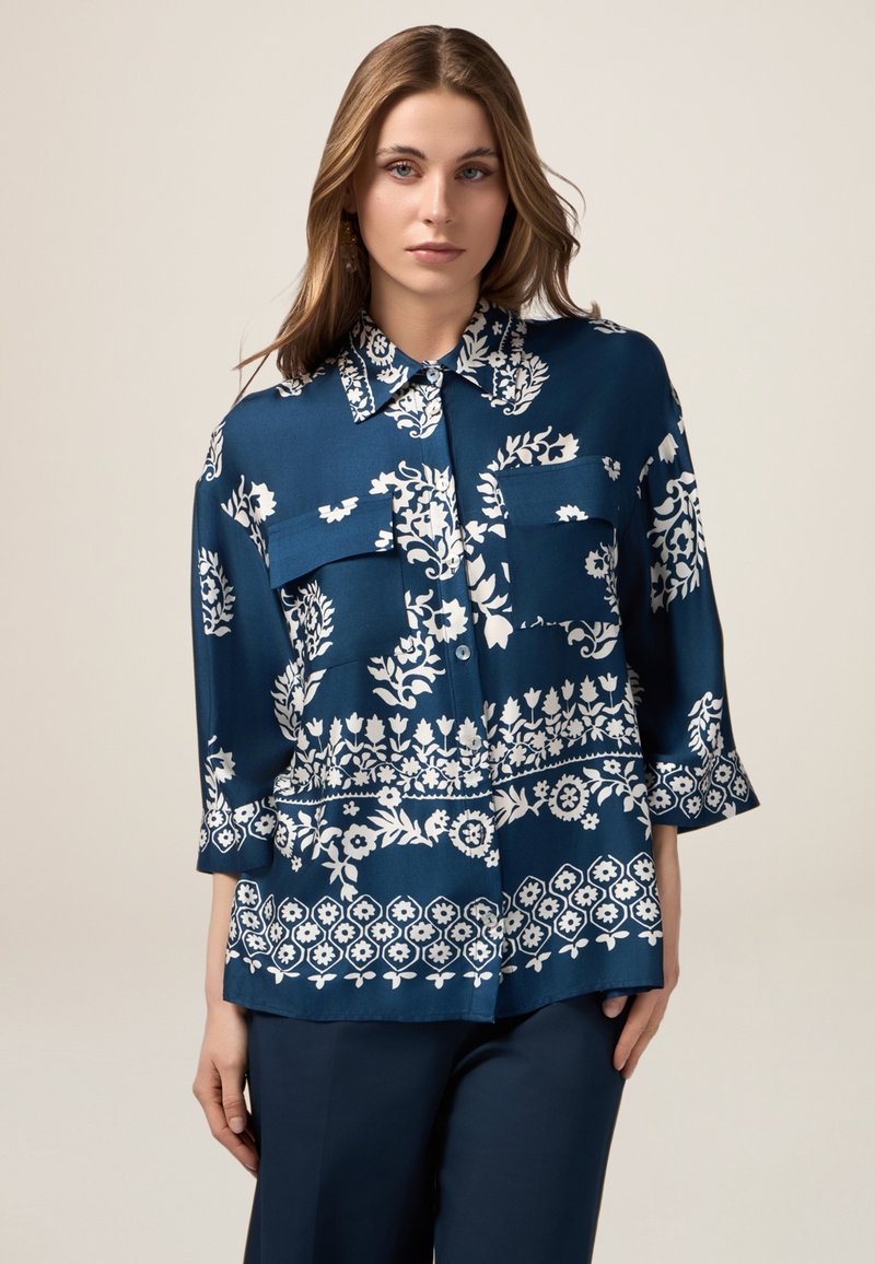 Blusa de tela suave con estampado floral azul, cuello tipo camisa, dos bolsillos frontales y mangas de 3/4.