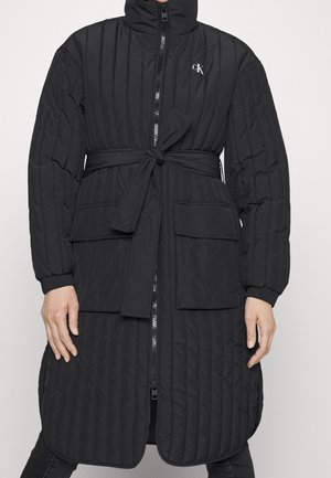 Long manteau noir matelassé avec col haut, fermeture éclair à l'avant, taille ceinturée, deux grandes poches à rabat et logo CK sur la poitrine.