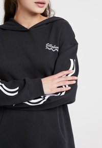 Sudadera negra con bolsillo frontal, logo blanco y patrones de olas en las mangas. Tejido suave, capucha ajustable y puños acanalados.