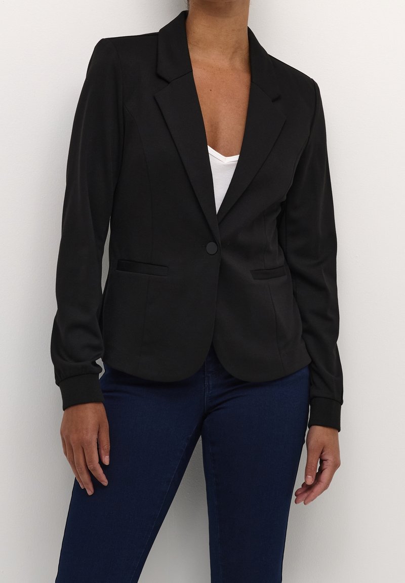 Zwarte blazer met een enkele knoopsluiting, twee voorzakken en een slanke pasvorm. Stevige stof met getextureerde mouwmanchetten, gedragen over een witte top.