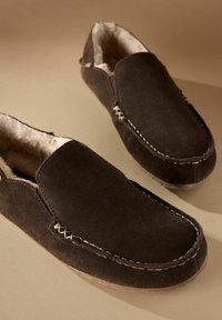Brune ruskind mokasin slippers med blød fleece-foring, elastiske sidepaneler og dekorativ syning på overdelen og sålen.