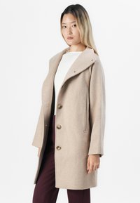 Femme portant un manteau beige jusqu'aux genoux avec de grands revers et trois boutons, associé à un haut blanc et un pantalon violet foncé sur fond uni.