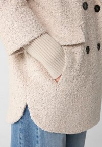 Hellbeige doppelt-knäpfiger Mantel aus strukturiertem, flauschigem Material. Verfügt über große schwarze Knöpfe und ein geripptes, gestricktes Bündchen, das aus dem Ärmel herausschaut.