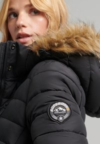 Sort pufferjakke med hætte trimmed med pels, quiltet design og et rundt logo-patch på venstre ærme.