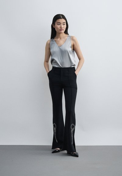 Γυναίκα στέκεται με τα χέρια στις τσέπες φορώντας ασημί αμάνικη μπλούζα, μαύρο παντελόνι με φαρδιά ράσπα και λευκή κεντητική, και μαύρα μυτερά παπούτσια.