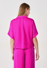 Blusa de satén rosa de manga corta con un corte holgado, que presenta un canesú en la parte posterior y una textura suave, combinada con pantalones a juego.