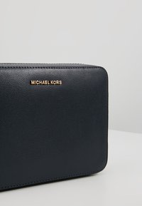 Černá kožená kabelka s texturovaným povrchem, zdobená zlatým logem "MICHAEL KORS" a zipovým uzavíráním, obdélníkového tvaru a čistými liniemi.