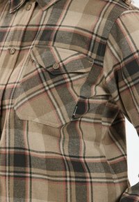 Bruine en zwarte geruite flanellen shirt met twee borstzakken, knoopsluitingen en een zachte textuur. Opmerkelijke rode accentlijnen.