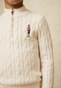 Pull en maille couleur crème avec un col haut et une demi-zip, présentant un petit dessin de casse-noisette brodé sur la poitrine.